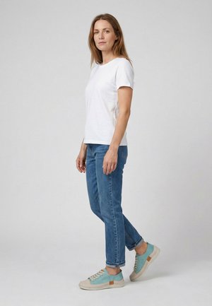 Kvinde stående iført en hvid kortærmet skjorte, blå jeans og blå/beige casual sneakers mod en ensfarvet grå baggrund.