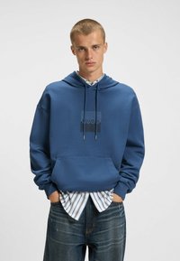 Felpa blu con cappuccio, dotata di tasca frontale e lacci. Presenta una grafica del logo ed è indossata sopra una camicia a righe bianca e blu.