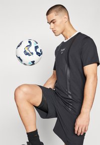 Zwart sport t-shirt met textuurstrepen, gecombineerd met zwarte shorts. Een wit en blauwe voetbal zweeft op knielengte ervoor.