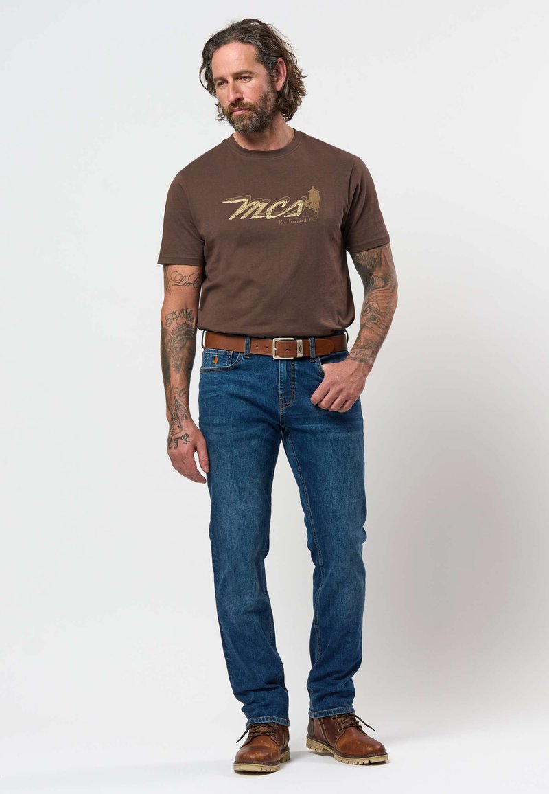 Bărbat cu tatuaje purtând un tricou maro "MCA", blugi albaștri, curea maro și cizme maro, stând în fața unui fundal simplu și luminos.