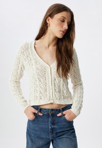 Strickjacke in Creme mit V-Ausschnitt, raffiniertem Spitzenmuster, Knopfverschluss und langen Ärmeln, kombiniert mit hochtaillierten blauen Jeans.