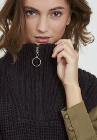 Mørk, chunky strikket sweater med høj krave, der har en sølv lynlås med en cirkulær træk. Glat olivengrøn manschet synlig.