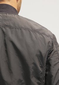 Grå letvægts bomberjakke med ribbet krave, glat overflade og syninger på skulder og ryg. Vandtæt materiale.