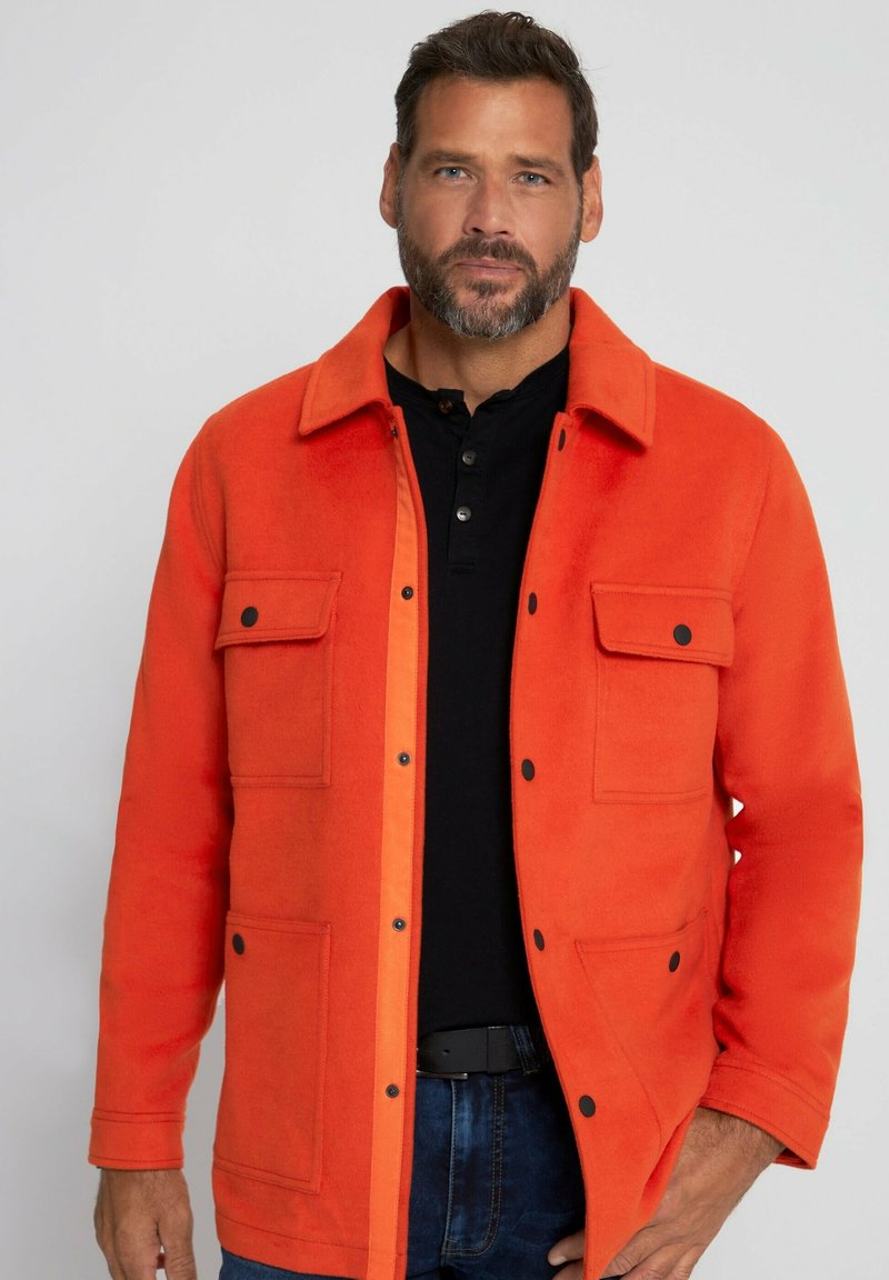 Oranger Fleece-Jacke mit Kragen, zwei Brusttaschen und schwarzen Knöpfen, getragen über einem schwarzen Hemd mit Knöpfen.