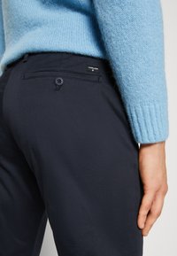 Pantaloni navy dal tessuto liscio, con una tasca posteriore dettagliata da un bottone e un'etichetta del marchio. Abbinati a un maglione azzurro chiaro.