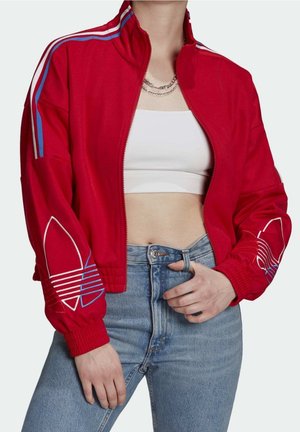 Personne portant une veste rouge zippée avec des rayures bleues et blanches, un crop top blanc, des colliers en chaîne superposés et un jean bleu taille haute.