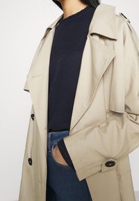 Beige Trenchcoat mit doppelter Knopfleiste, großen Reverskragen und schwarzen Knöpfen, getragen über einem marineblauen Oberteil und blauen Jeans.