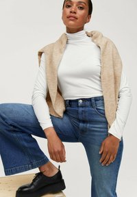 Cremefarvet turtleneck top under en beige ærmeløs cardigan, parret med højtaljede blå jeans og sorte snørebåndssko. Teksturerede stoffer synlige.