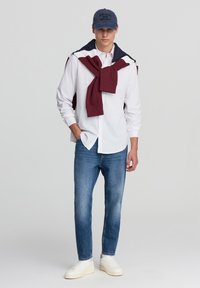 Biała koszula z guzikami, niebieskie dżinsy, białe trampki. Bordowy sweter zarzucony na ramiona. Ciemnoniebieska czapka z napisem. Prosta, casualowa stylizacja.