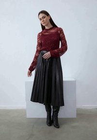 Burgundi csipkés, hosszú ujjú felső fekete pliszírozott midi szoknyával párosítva. A modell fekete térd fölé érő csizmát visel. Semleges háttér világos padlóval.
