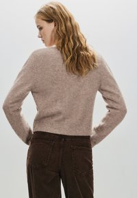 Femeie cu păr blond ondulat, purtând un pulover bej tricotat și pantaloni maro închis din catifea, stând cu mâinile în șolduri, cu spatele la cameră.