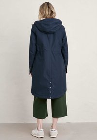 Marineblauwe waterdichte jas met capuchon, knielange ontwerp, knoopdetails aan de achterkant, gecombineerd met groene cropped pants en witte sneakers.