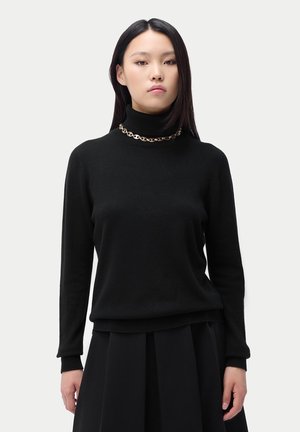 Jeune femme aux longs cheveux foncés portant un pull à col roulé noir, un collier chaîne en or et une jupe plissée noire, debout devant un fond uni.