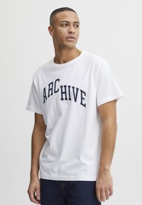 Bílá bavlněná tričko s krátkým rukávem, s námořnickým zakřiveným textovým designem, který říká "ARCHIVE" přes hruď. Jednoduchý, uvolněný střih.