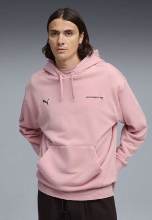 Joven con cabello oscuro hasta los hombros que lleva una sudadera con capucha Puma x Porsche de color rosa claro y pantalones negros, sobre un fondo gris.