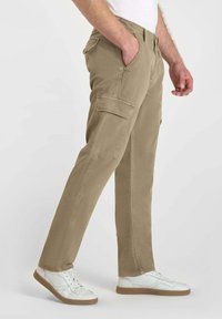 Man som bär beige cargobyxor med sidofickor och vita sneakers, stående med en hand i fickan mot en enkel bakgrund.