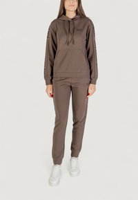 Mujer vistiendo una sudadera marrón de EA7 y pantalones de chándal a juego, combinados con zapatillas blancas, de pie contra un fondo claro y liso.