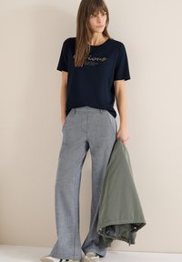 Femme portant un t-shirt graphique bleu marine et un pantalon large gris, tenant une veste verte, debout contre un mur beige.