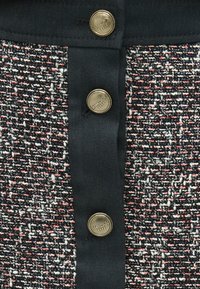 Melnas un krāsainas tweeda audums ar teksturētu rakstu, ar zelta pogas aizdari un melnu apmali gar centru.