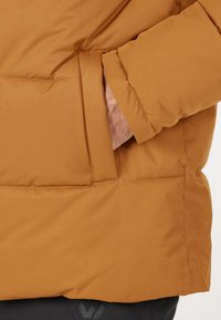 Brun puffjacka med en slät textur, med en sidoficka och minimal sömnad. Den figursydda manschetten visar en hand inuti.