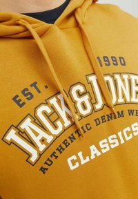 Felpa gialla con cappuccio, caratterizzata da una grafica con logo frontale contenente le scritte "JACK & JONES" e "CLASSICS" in bianco e navy. Include un cappuccio regolabile con cordini.