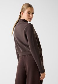 Vrouw met gevlochten haar, gekleed in een lange mouw bruine top en bijpassende broek, die over haar schouder kijkt tegen een effen achtergrond.