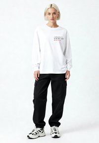 Giovane donna con capelli biondi corti indossa una maglietta bianca a maniche lunghe con testo e pantaloni cargo neri, in piedi davanti a uno sfondo semplice.