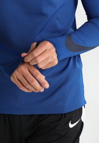 Maglietta sportiva a maniche lunghe in tessuto blu con un finish testurizzato. Presenta un accento blu scuro sulla manica e un orlo arrotondato. Pantaloni corti neri.