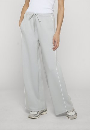 Person iført lysegrå vidde-sweatpants med hvid piping, hvid tanktop og grå-hvide sneakers, stående med hænderne i lommerne.