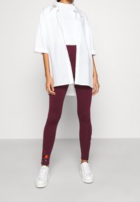 Burgundy leggings feature floral accents at the ankles, paired with a white, oversized button-up shirt and white sneakers.