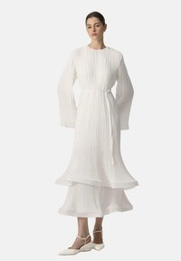 Robe blanche plissée avec des manches longues, taille cintrée et ourlet à volants. Le mannequin porte des chaussures plates blanches à bouts pointus avec des brides à la cheville.