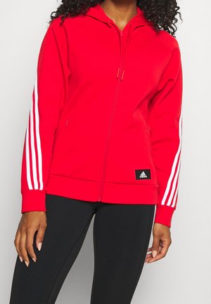 Kobieta ubrana w czerwoną bluzę Adidas na zamek z białymi paskami na rękawach oraz czarne legginsy z bocznymi paskami.