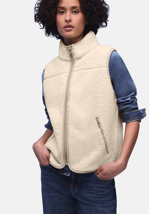 Donna con capelli ricci che indossa un gilet in pile color crema con cerniera sopra una camicia di jeans arrotolata e jeans blu scuro, mani nelle tasche.