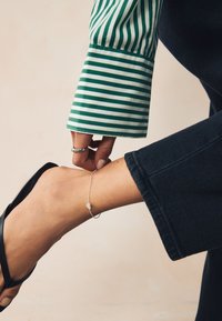 Schwarze Flip-Flop-Sandale, am Knöchel geschmückt mit einem zarten silbernen Kettenarmband mit einem kleinen Herzanhänger, kombiniert mit dunkler Denim und einem grün gestreiften Ärmel.