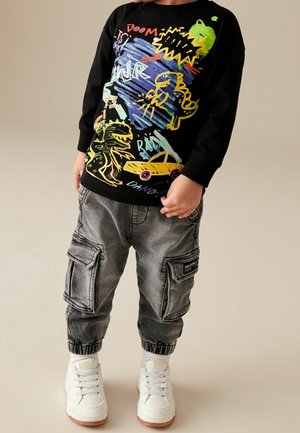 Enfant portant un sweat-shirt noir avec un graffiti de dinosaure coloré et de voiture, un pantalon cargo gris et des baskets blanches sur fond beige.