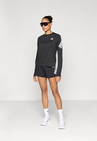 Čierne športové tričko s dlhým rukávom a bielymi pruhmi, doplnené čiernymi šortkami. Model má na sebe čierne slnečné okuliare a biele tenisky s jemnými detailmi.