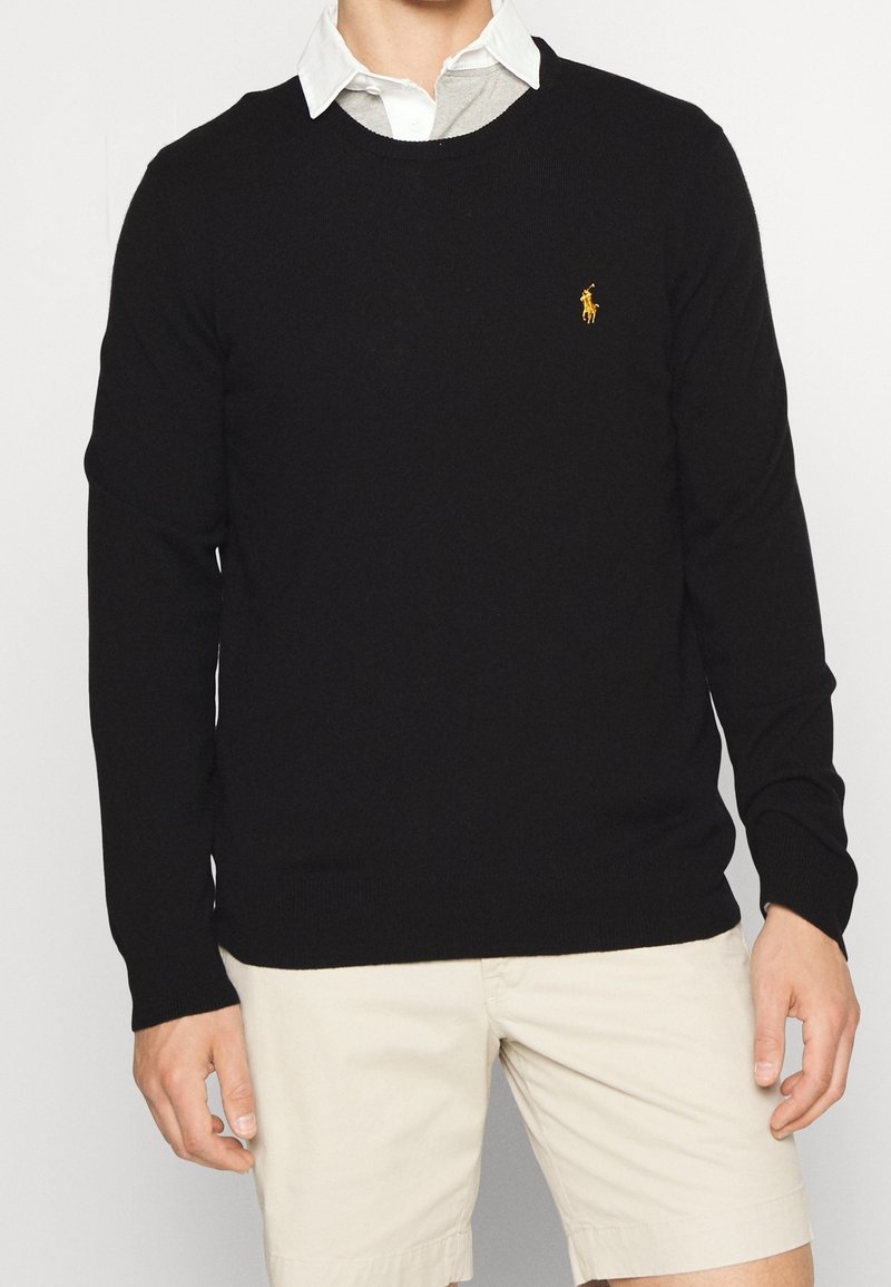 Homme portant un pull noir à manches longues avec un petit logo jaune sur la poitrine, une chemise blanche à col et un short beige sur fond clair.