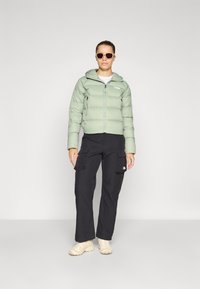 Lichtgroene pufferjack met capuchon, zwarte cargo-broek en beige schoenen. Model met zonnebril, staande tegen een effen achtergrond.