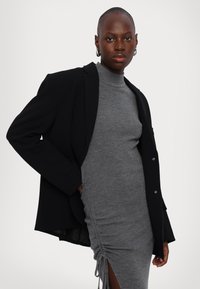 Mustapukuiset blazerit harmaan ribbyn turtleneck -mekon päällä, jossa on sivu rypytys ja halkio, esittelee sulavaa siluettia ja tasaisia tekstuureja.