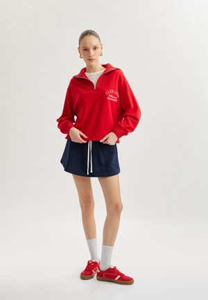 Rød zip-up sweatshirt med ribbet krave, parret med en marineblå trækbånds nederdel og røde sneakers, der har hvide accenter og grå sokker.