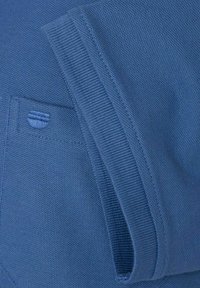 Niebieska dziana tkanina o fakturowanej powierzchni. Zawiera małą naszywkę i zamek błyskawiczny wzdłuż krawędzi, z prążkowanymi mankietami i dołem.