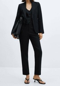 Completo tre pezzi nero composto da un blazer aderente, un gilet con bottoni e pantaloni a gamba dritta, abbinato a sandali con tacco a flip-flop neri e una borsa a mano.