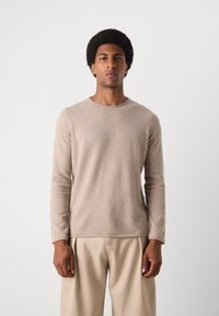 Beige Pullover mit langen Ärmeln, rundem Ausschnitt, glatter Textur und subtilen Muster, kombiniert mit hellen, plissierten Stoffhosen.