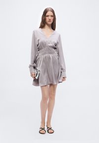 Γυναίκα που φοράει μακρυμάνικο φόρεμα σε ανοιχτό μωβ με V λαιμόκοψη και ζώνη στη μέση, κρατώντας ασημένιο τσαντάκι χειρός και φορώντας μαύρα σανδάλια, στέκεται πάνω σε λευκό φόντο.