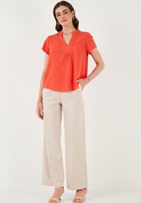 Blusa corta arancione brillante con scollo a V e cuciture decorative sottili. Abbinata a pantaloni beige larghi e sandali di colore chiaro.