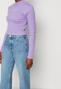 Osoba nosící přiléhavý lila top s dlouhým rukávem a světlé modré džíny s vysokým pasem na jednobarevném pozadí.