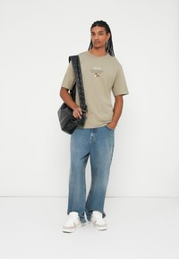 T-shirt kaki avec un motif imprimé, associé à un jean bleu ample et des baskets blanches. Un sac noir avec des détails cloutés est porté.