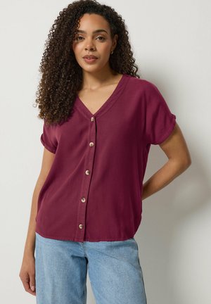 Frau mit lockigem Haar, die eine kurzärmelige bordeauxrote Bluse mit Knöpfen und hellblaue Jeans trägt, steht vor einer weißen Wand.