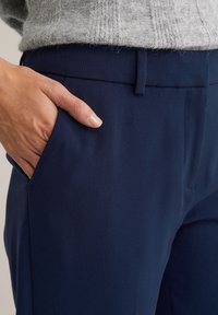 Pantalones ajustados azul marino hechos de una tela texturizada, con un bolsillo lateral y una cintura estrecha, con una mano colocada de manera casual en el bolsillo.