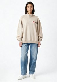 Sweat-shirt beige zippé avec une broderie « I ❤️ you » en rouge et noir, associé à un jean large bleu clair et des baskets blanches.